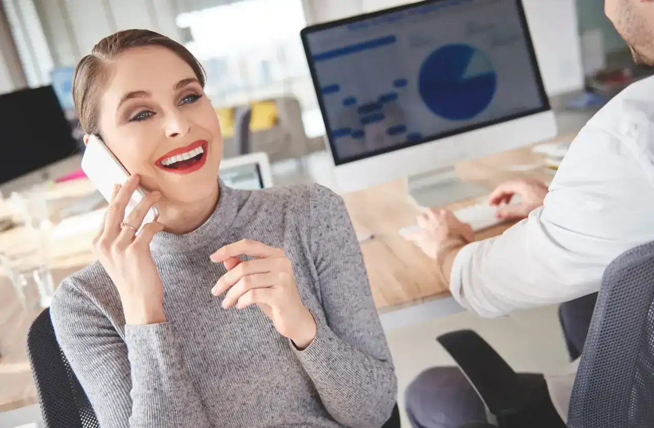 Uśmiechnięta kobieta rozmawia przez telefon, pytając, czy umowa w Play przedłuża się automatycznie. Obok mężczyzna pracuje przy komputerze.