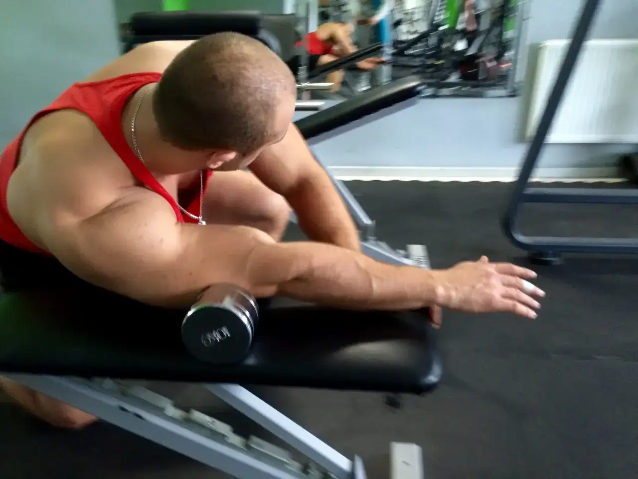 Mężczyzna w czerwonej koszulce ćwiczy przedramię z hantlem na ławce w siłowni.