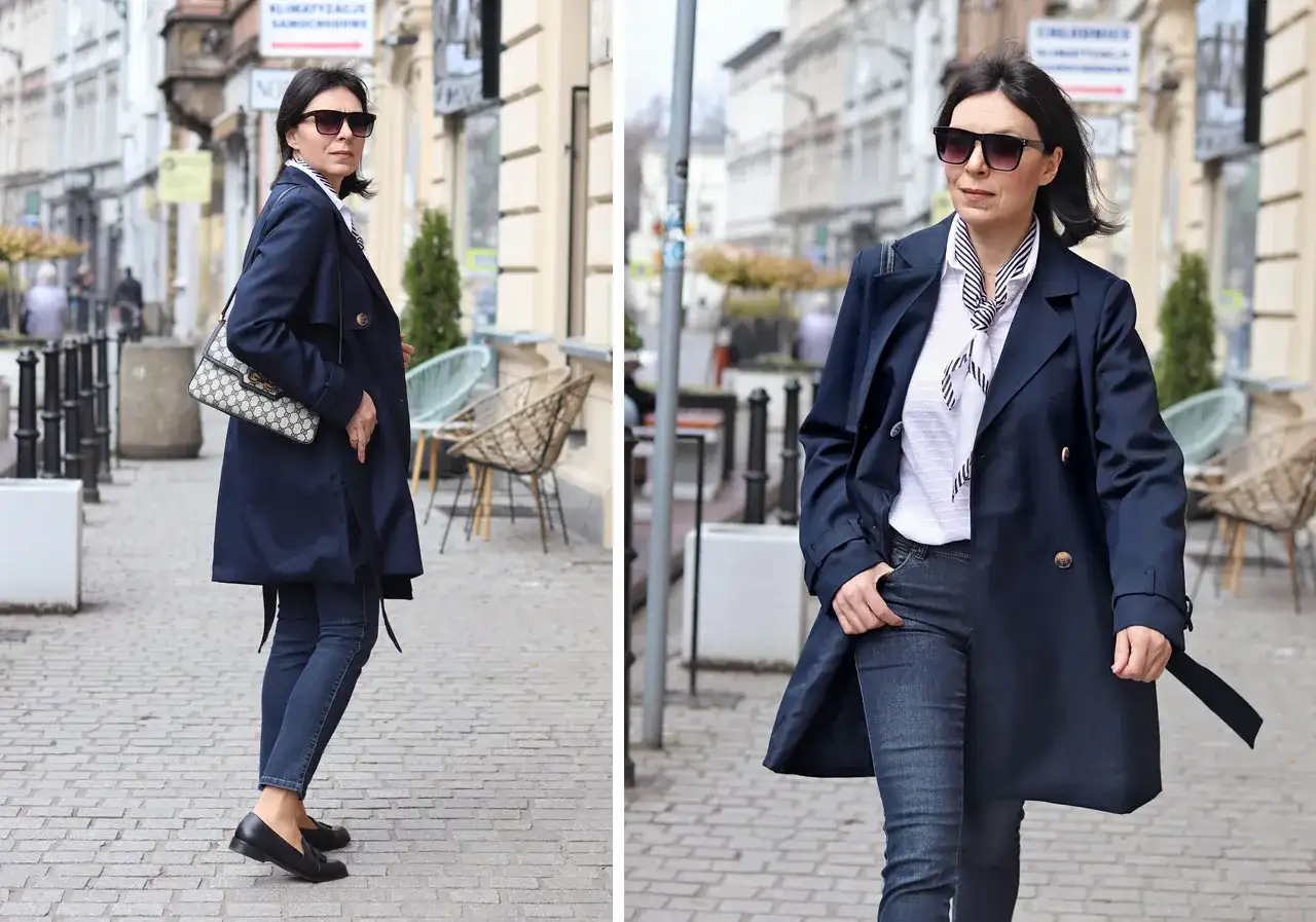 Elegancka kobieta w granatowym trenczu damskim, białej bluzce w paski i jeansach spaceruje po brukowanej ulicy.