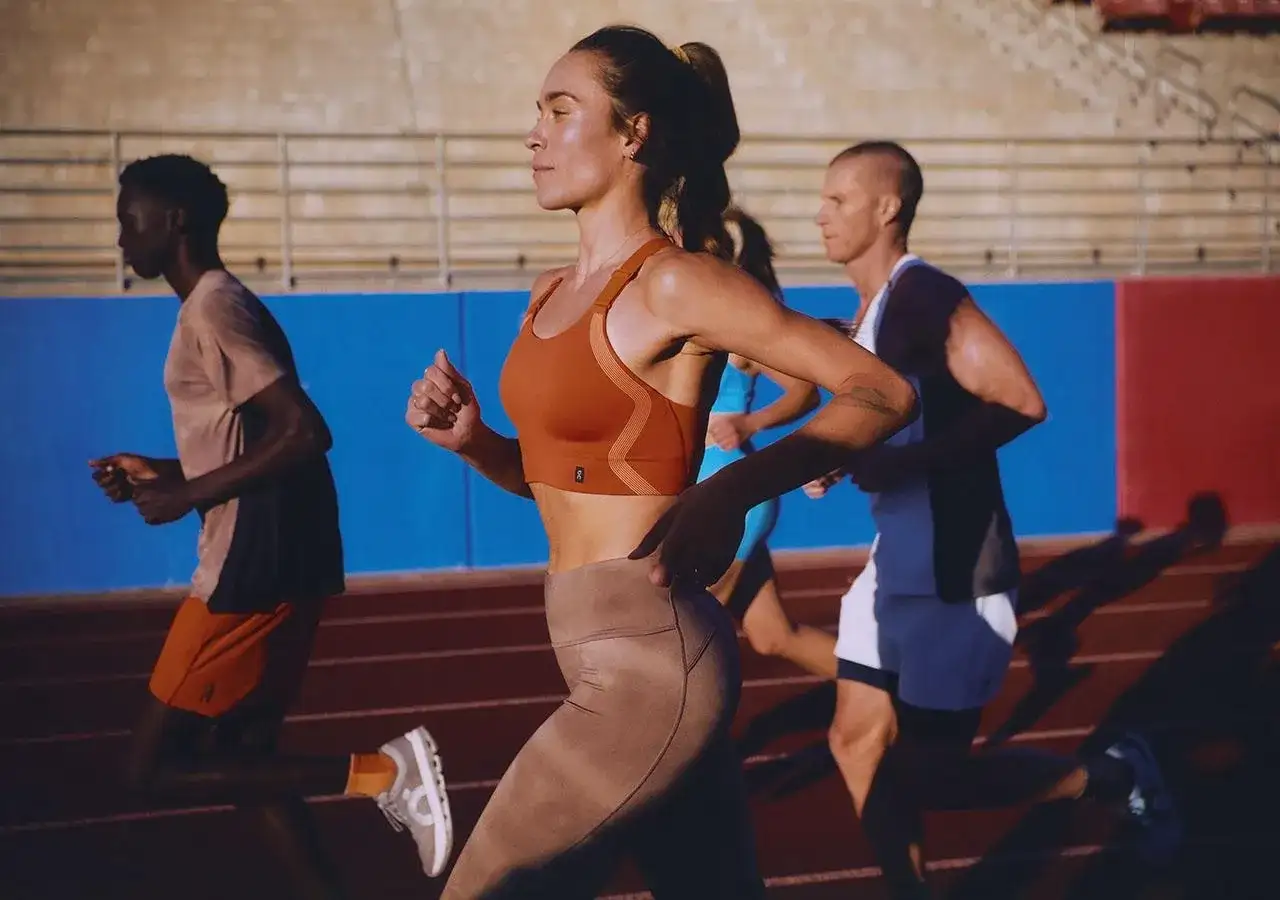 Trzech biegaczy na bieżni. Kobieta w pomarańczowym topie i beżowych legginsach, mężczyzna w beżowej koszulce i pomarańczowych spodenkach, oraz mężczyzna w białej koszulce i białych spodenkach. To jest ich "drop" formy.
