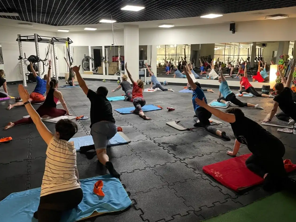 Grupa osób ćwiczy jogę na macie w siłowni Tychy Stadion. Lustra odbijają salę pełną ćwiczących.