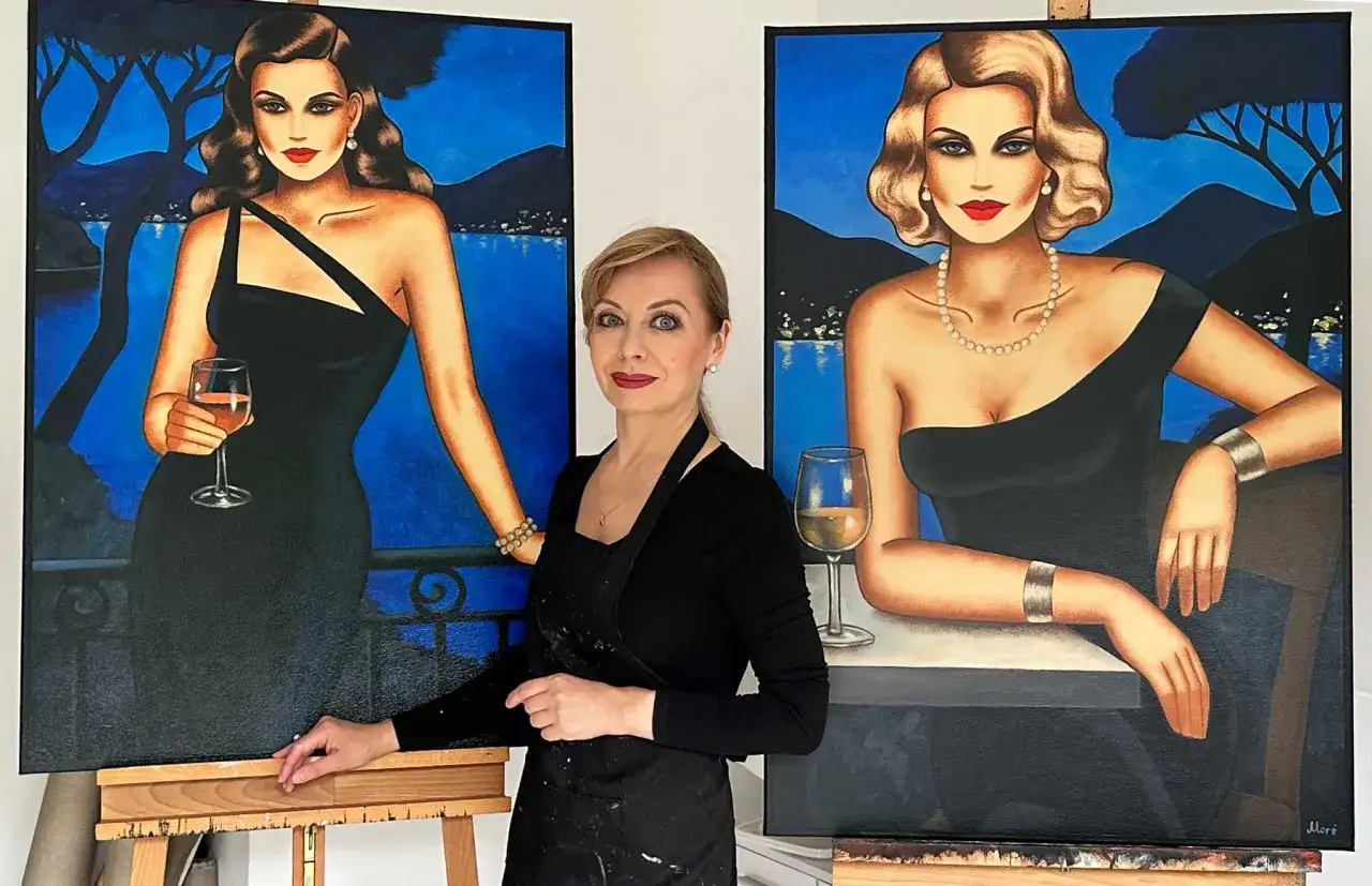 Künstlerin steht zwischen zwei Gemälden von eleganten Frauen in schwarzen Kleidern mit Gläsern Wein.
