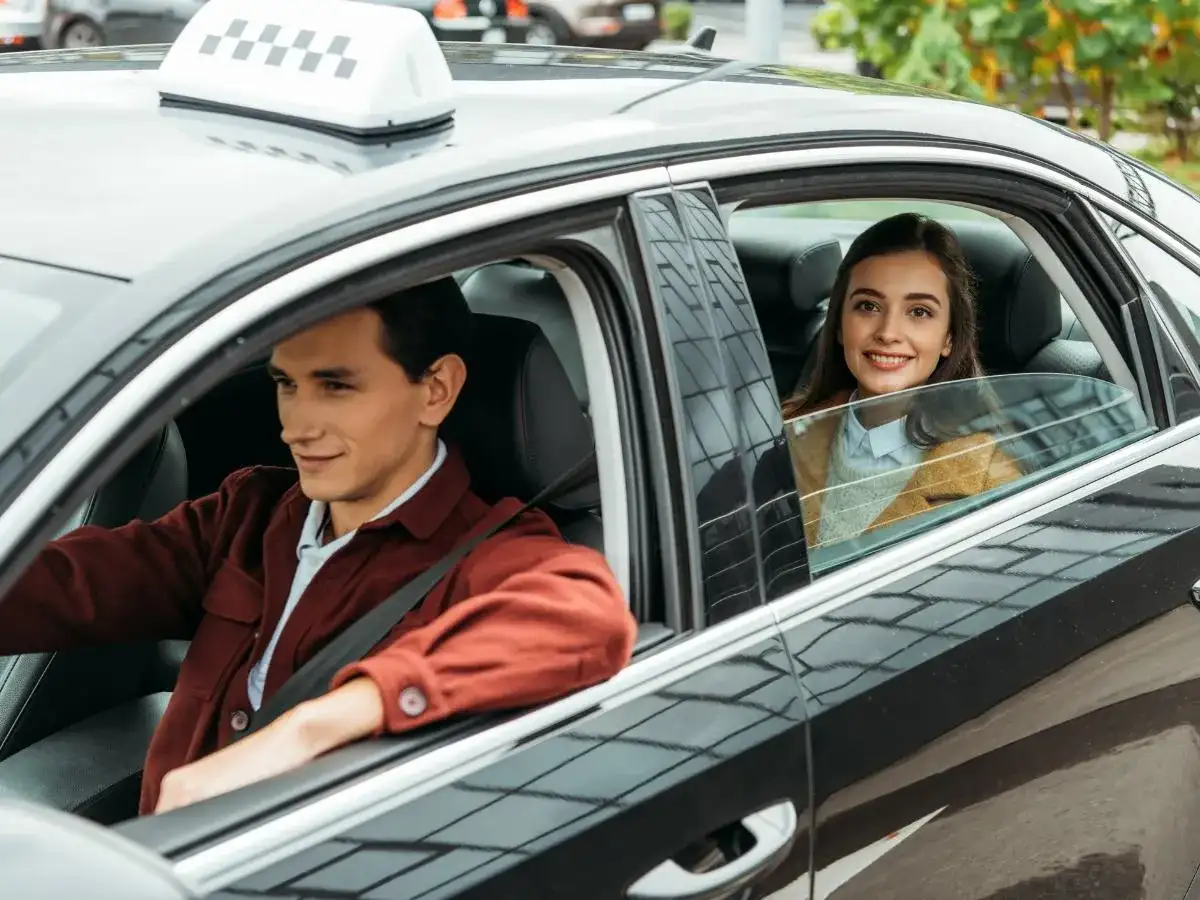 Kierowca taksówki i uśmiechnięta pasażerka. Czy pasażer taksówki ma obowiązek zapinania pasów bezpieczeństwa? Warto pamiętać o bezpieczeństwie.