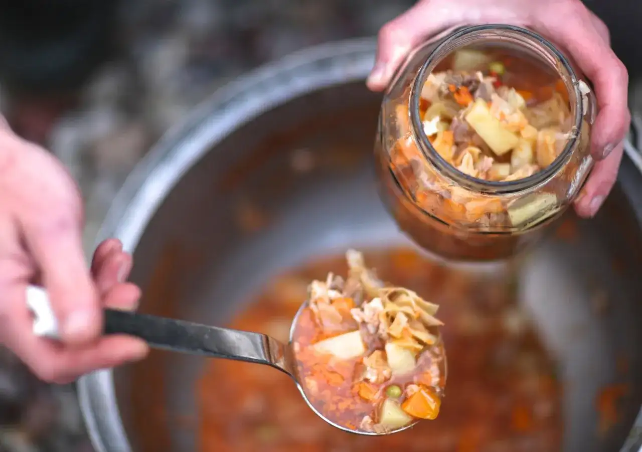 Łyżka zupy z warzywami i makaronem nad słoikiem, w tle garnek z zupą.