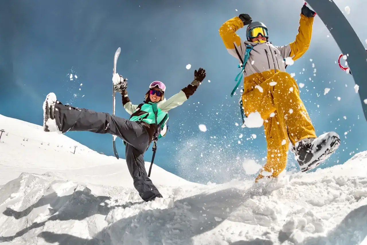 Dwóch narciarzy w akcji na śniegu, jeden z nartami, drugi na snowboardzie, z rozbryzgami śniegu.