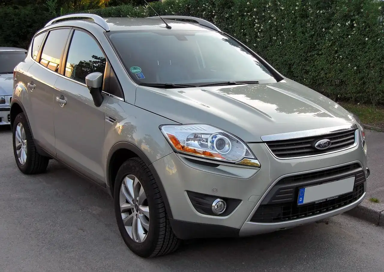 Szary Ford Kuga, SUV, prezentuje swoje wymiary na tle zielonego żywopłotu.