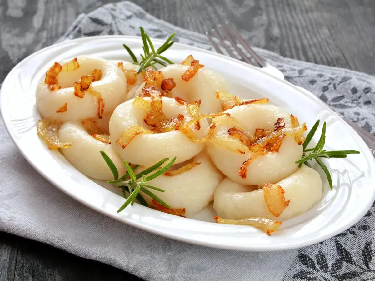 Kluski śląskie z podsmażoną cebulką i rozmarynem na białym talerzu.