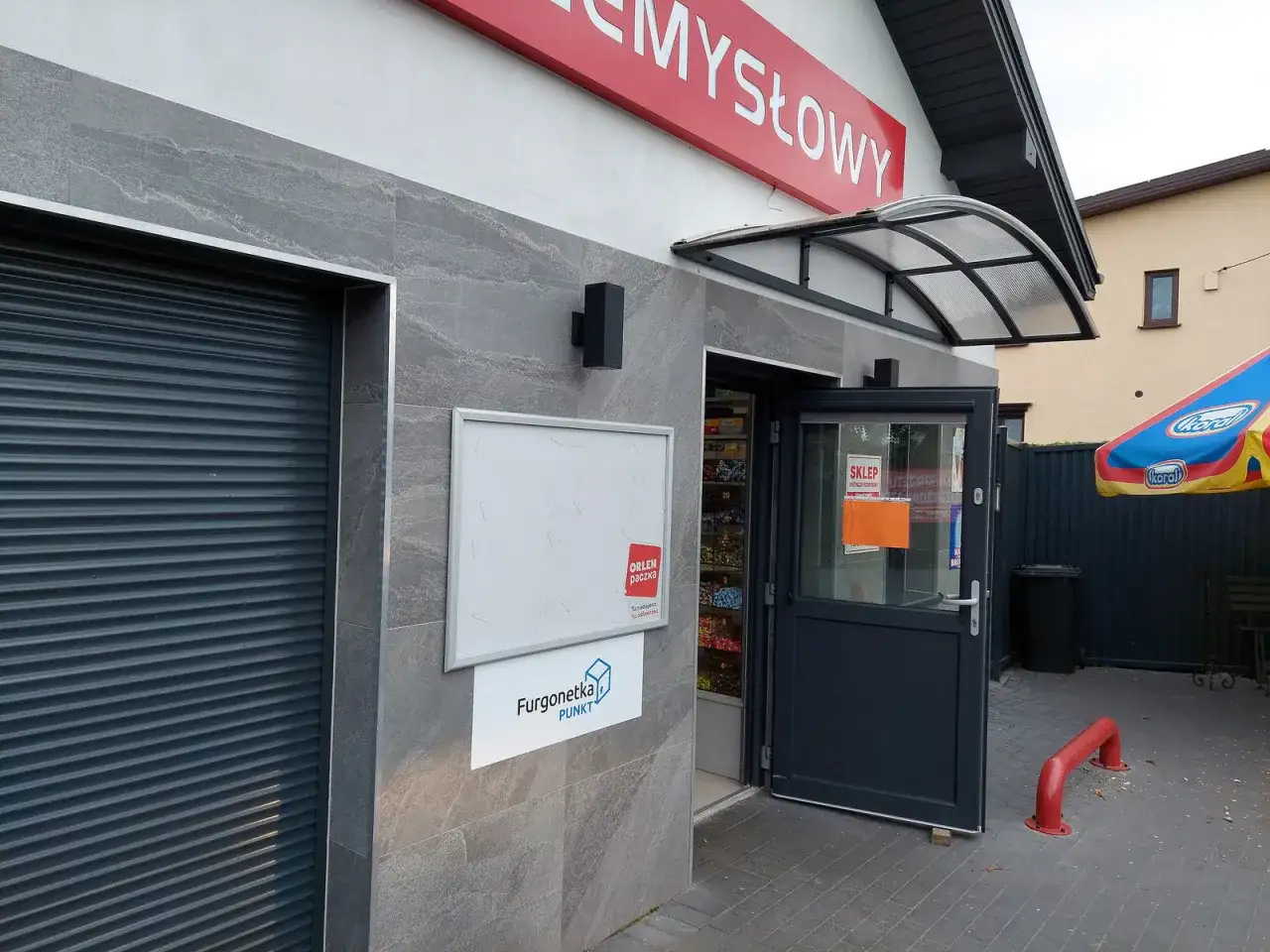 Wejście do sklepu z otwartymi drzwiami, tablicą ogłoszeń i logo "Orlen paczka".