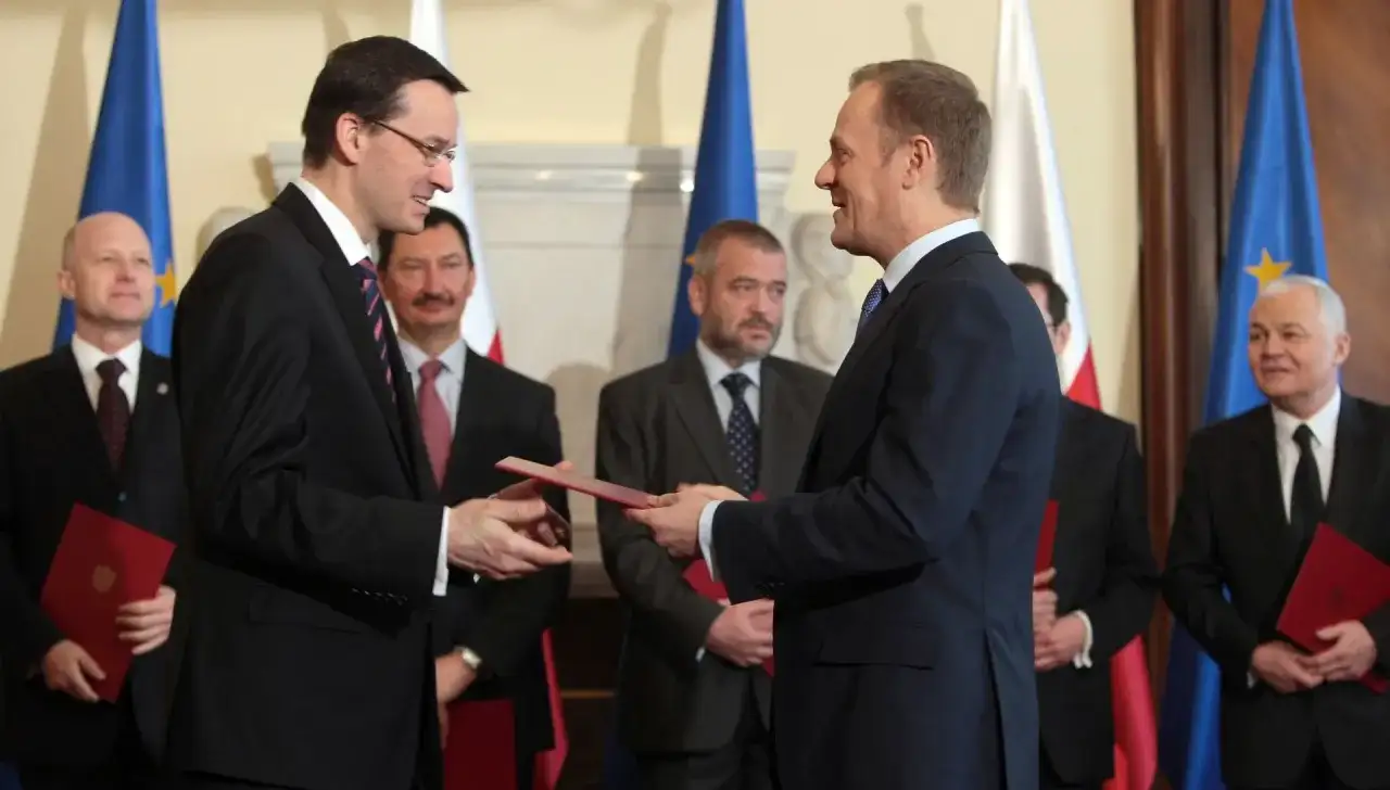 Mateusz Morawiecki przekazuje dokument Donaldowi Tuskowi, stojąc przed polskimi i unijnymi flagami.
