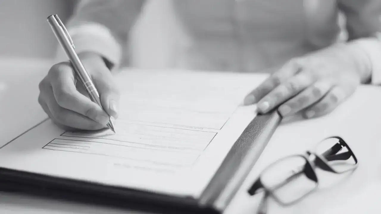 Dłoń pisze na dokumencie, obok okulary. Dowiedz się, jak sprawdzić pozwolenie na budowę sąsiada, analizując formalności.