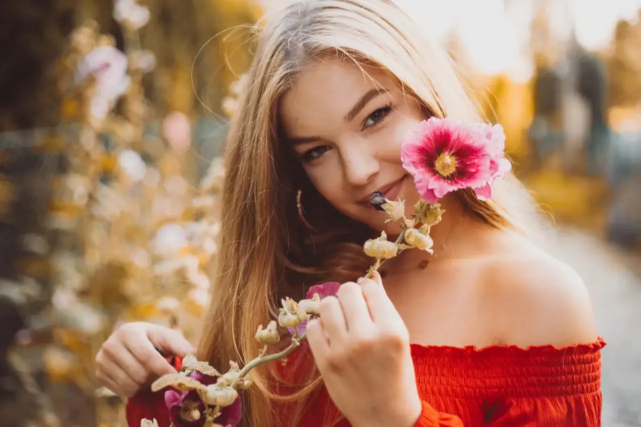 Uśmiechnięta blondynka w czerwonej bluzce podczas sesji kobiecej w plenerze, trzymająca gałązkę z różowymi kwiatami malwy.