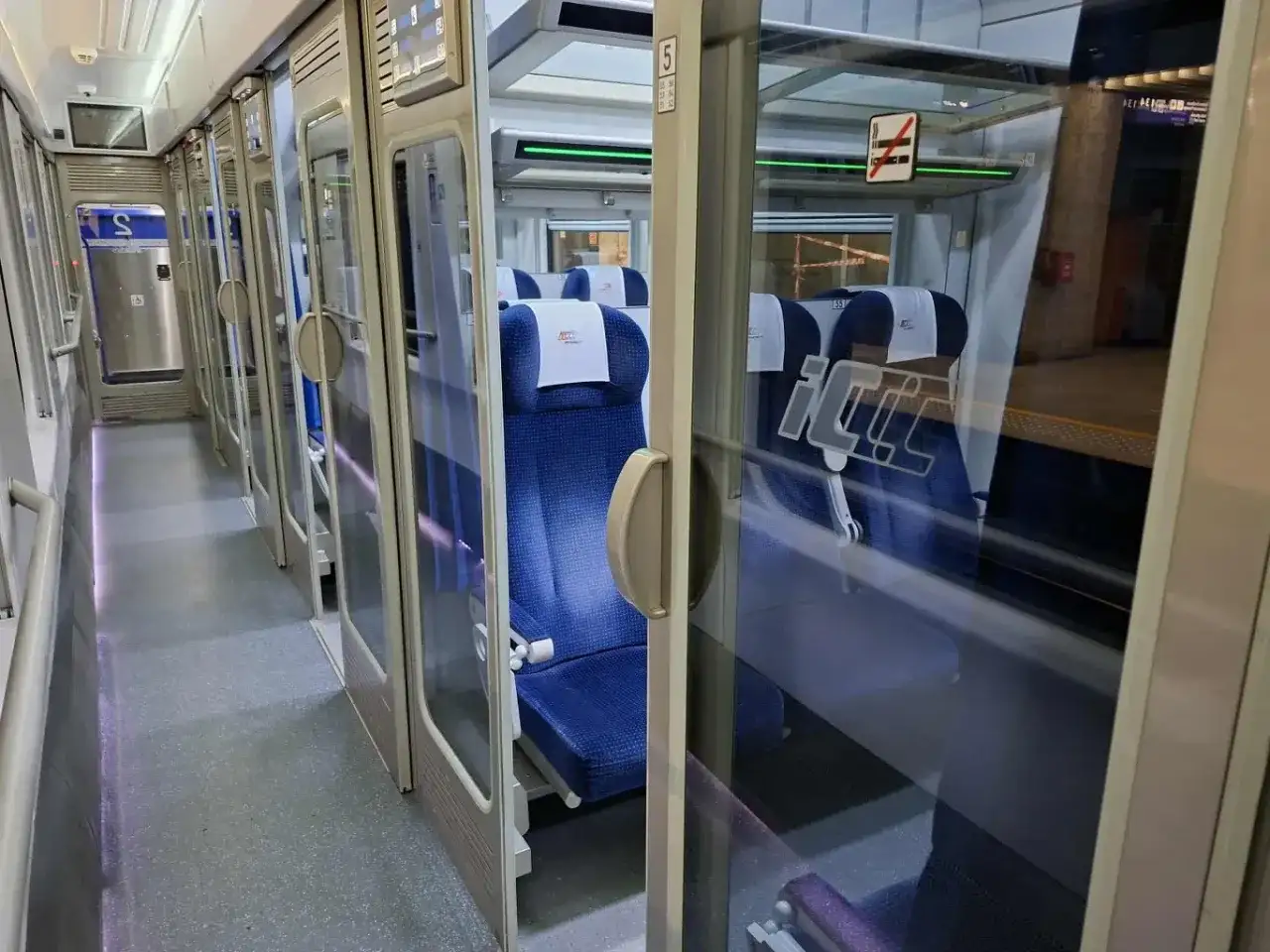 Wnętrze pociągu Intercity. Widać rząd niebieskich siedzeń i drzwi. Na szybie znak zakazu palenia, więc nie ma palarni.