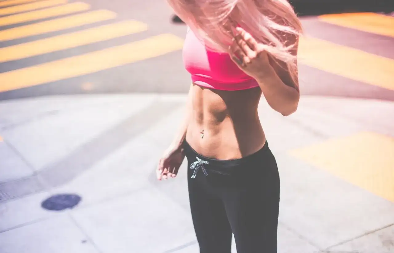 Kobieta z różowymi włosami w sportowym topie i legginsach prezentuje wyrzeźbiony brzuch.