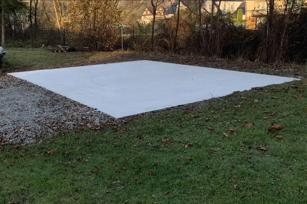 Nowa, jasna wylewka betonowa na gruncie, otoczona żwirem i trawą.