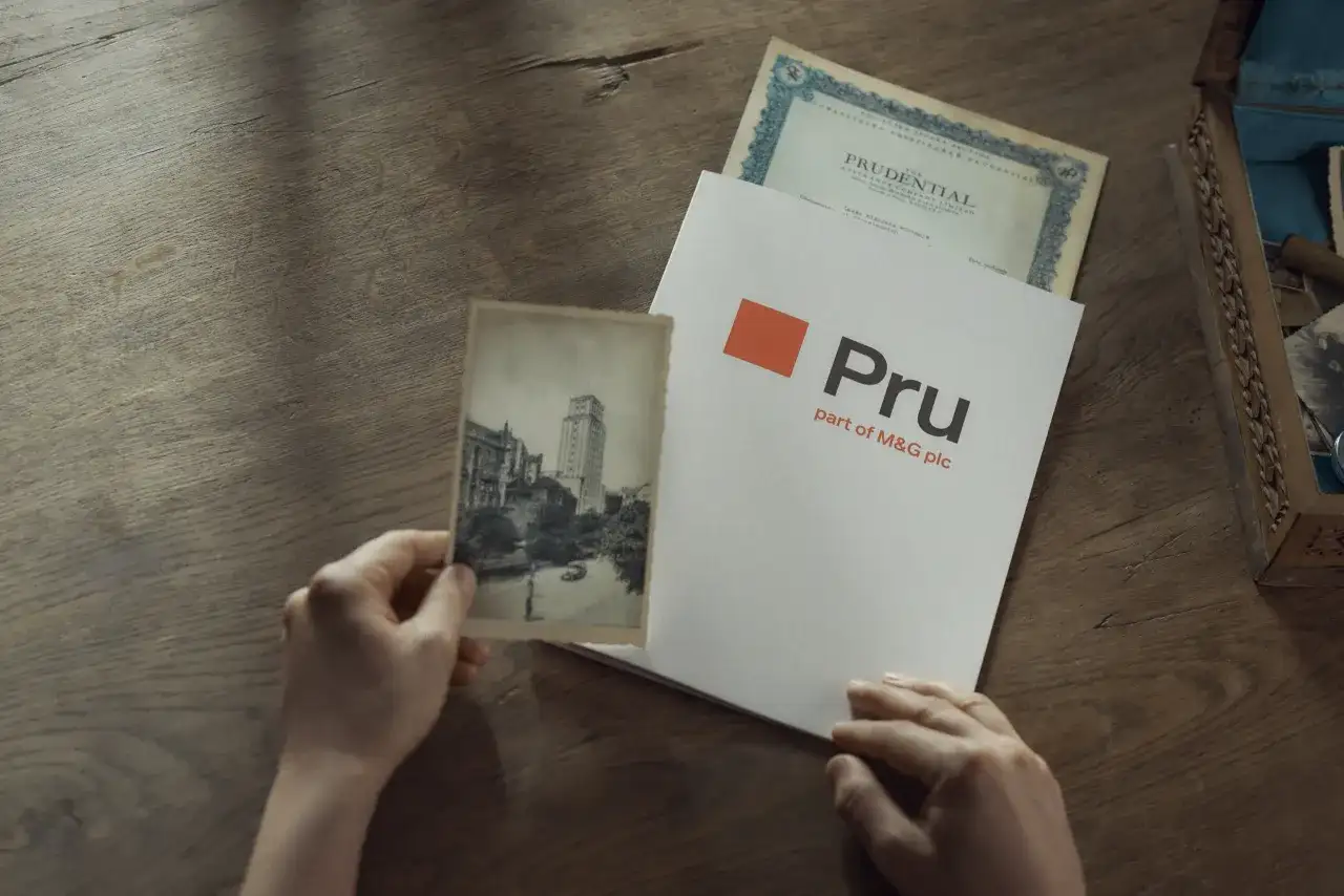 Ręce trzymają stare zdjęcie i dokument z logo Pru, part of M&G plc. To pokazuje, że Prudential to firma z długą historią.