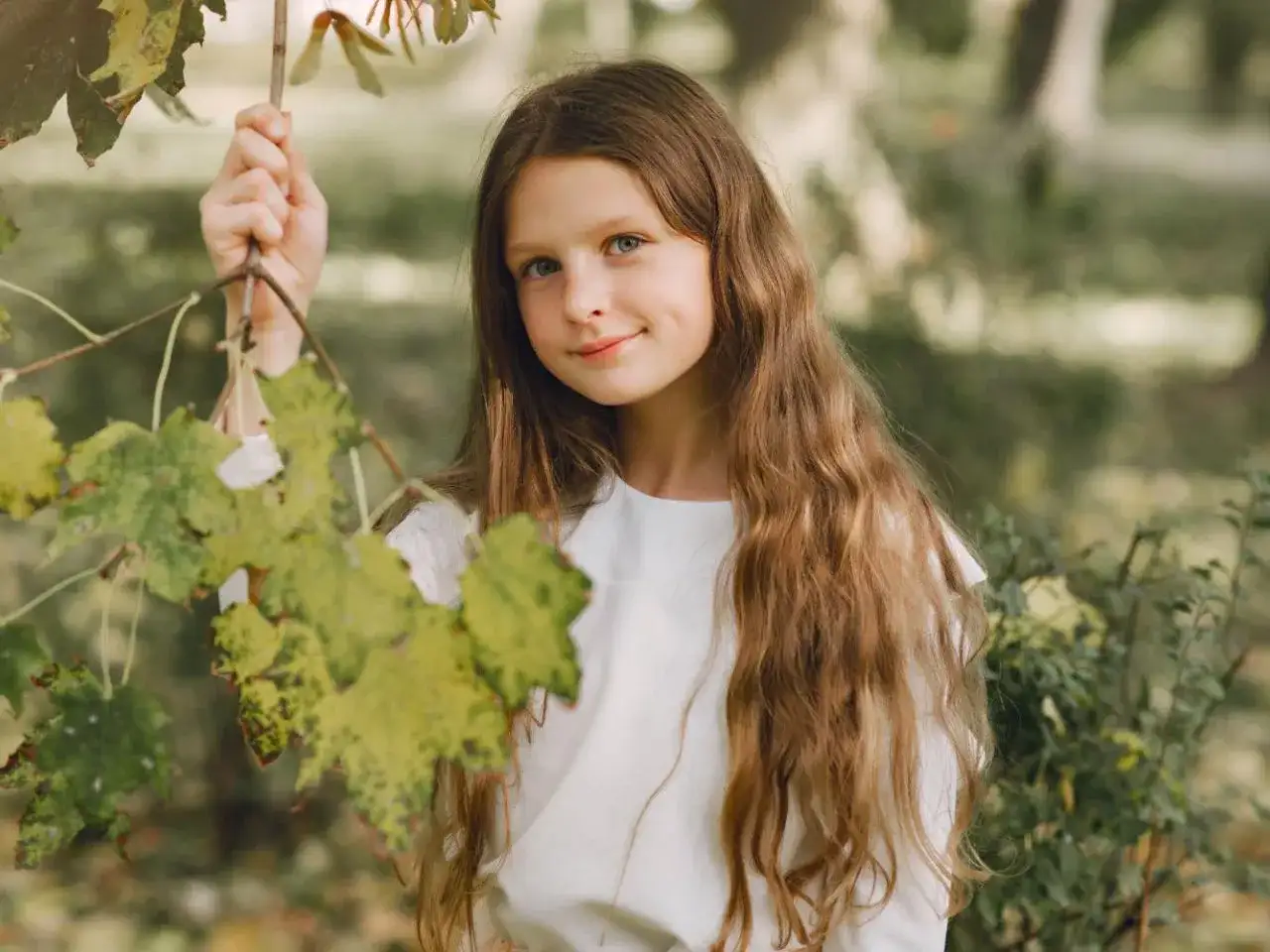 Dziewczynka z długimi włosami w białej bluzce trzyma gałązkę z liśćmi.