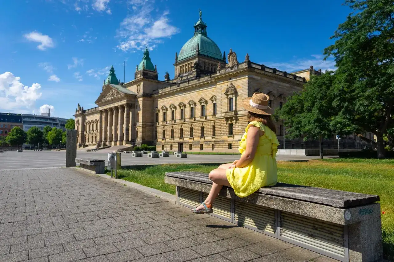 Kobieta w słomkowym kapeluszu i żółtej sukience siedzi na ławce przed monumentalnym budynkiem w Lipsku, mieście w Saksonii znanym z międzynarodowych targów.