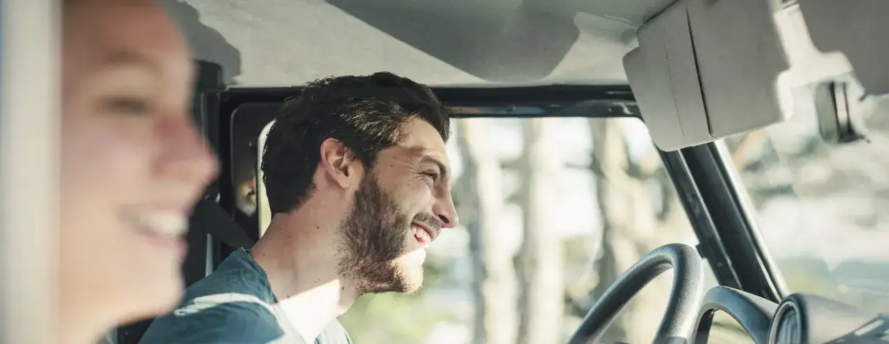 Uśmiechnięty mężczyzna za kierownicą, obok pasażerka. Planują podróż, myśląc o ubezpieczeniu auta na wyjazd za granicę.