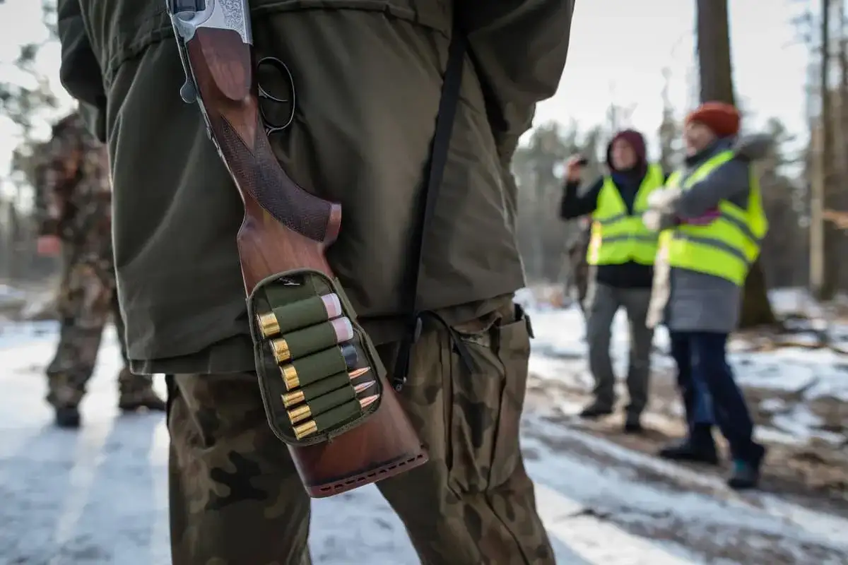 Myśliwy w lesie, z bronią i amunicją w pasie. W tle inni uczestnicy polowania w kamizelkach odblaskowych.