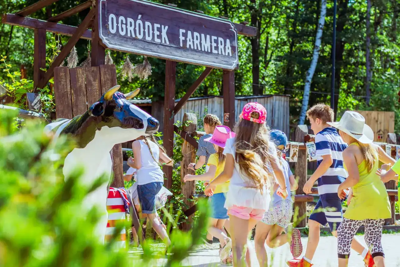 Dzieci biegną obok drewnianego szyldu "Ogródek Farmera" i figury krowy.