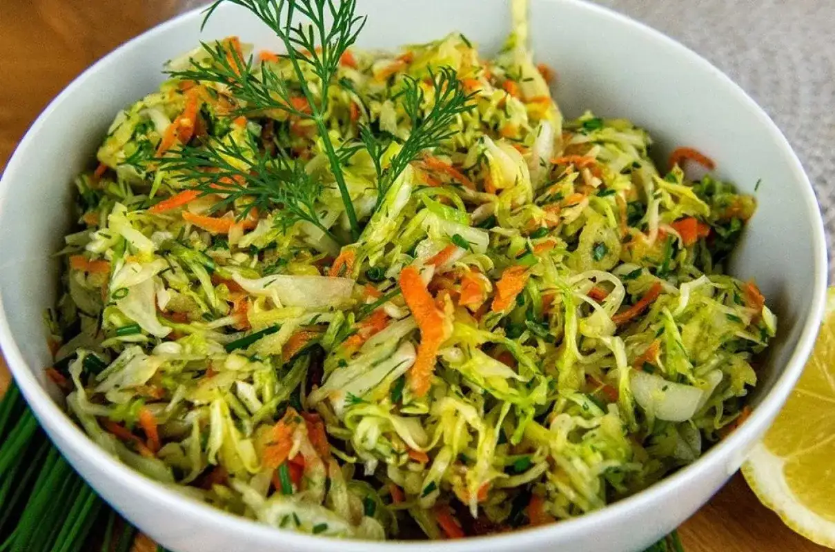 Surówka z kapusty i marchewki z koperkiem w białej misce, obok plasterek cytryny.