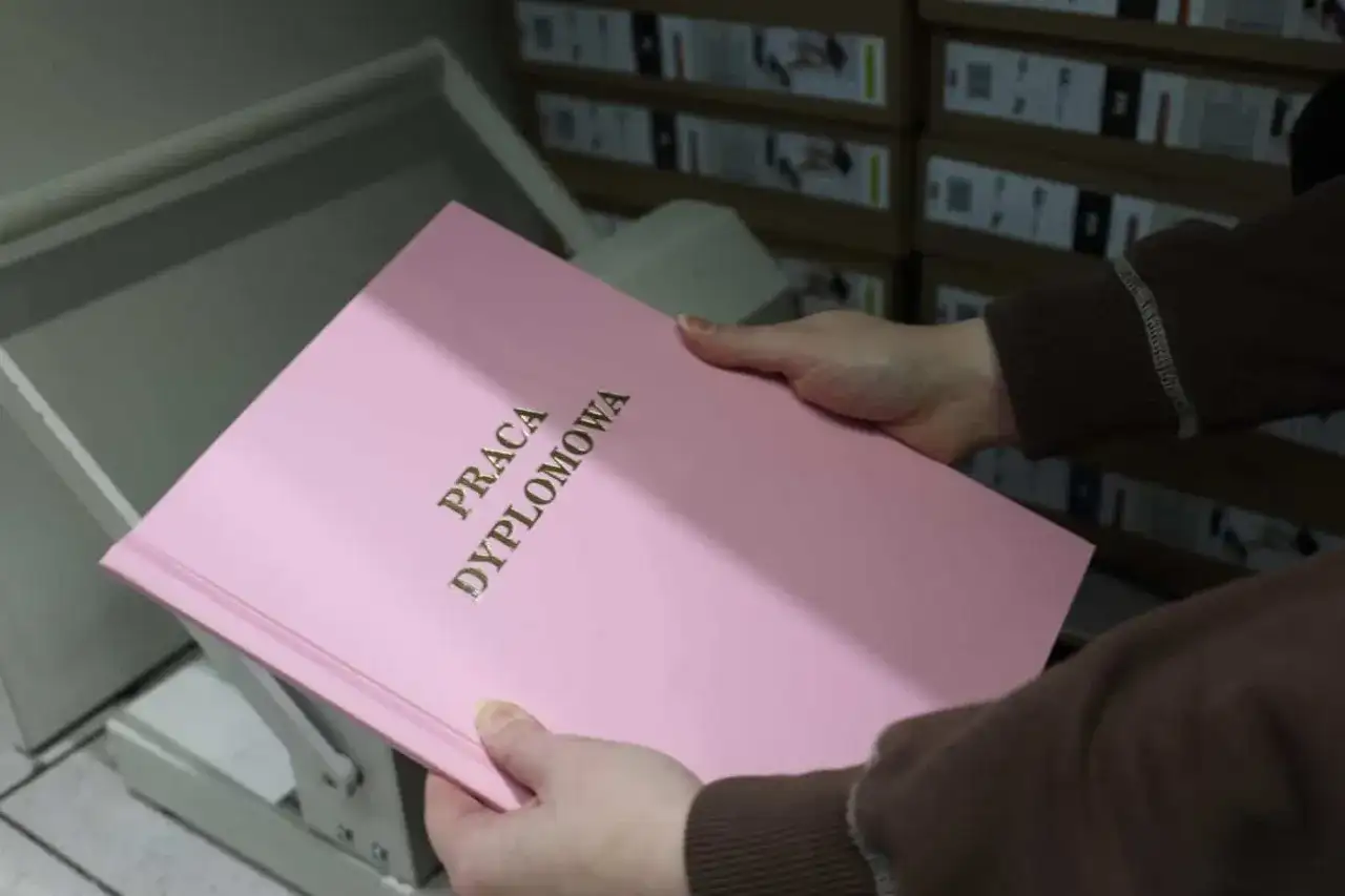 Różowa okładka pracy dyplomowej z napisem "PRACA DYPLOMOWA" w złoconych literach. W tle stosy pudeł, które mogą zawierać materiały do prezentacji.