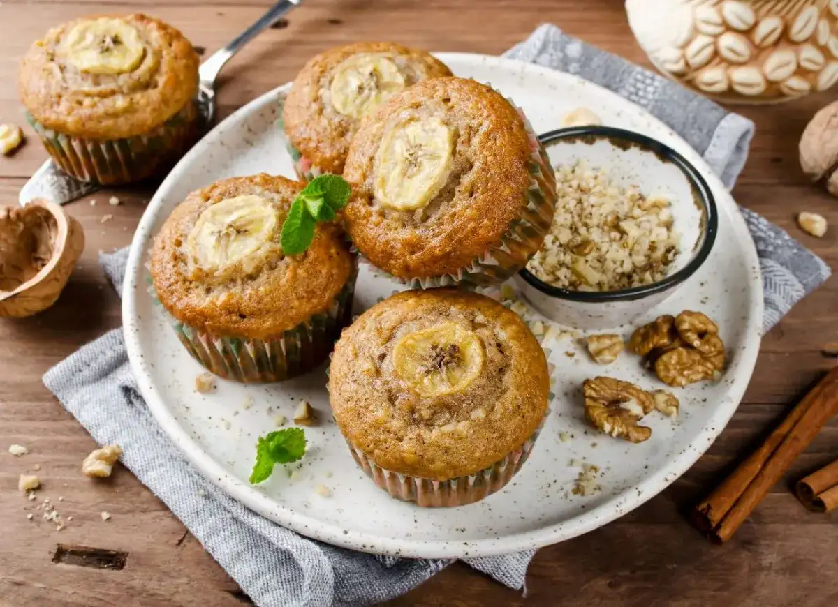 Pięć pysznych muffinek bananowych z plasterkiem banana na wierzchu, udekorowane miętą. Obok miseczka z orzechami i laska cynamonu.