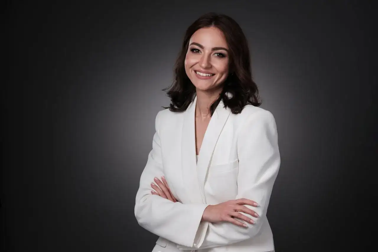 Uśmiechnięta kobieta w białej marynarce, z rękami skrzyżowanymi na piersi. Idealna sesja biznesowa pozy.