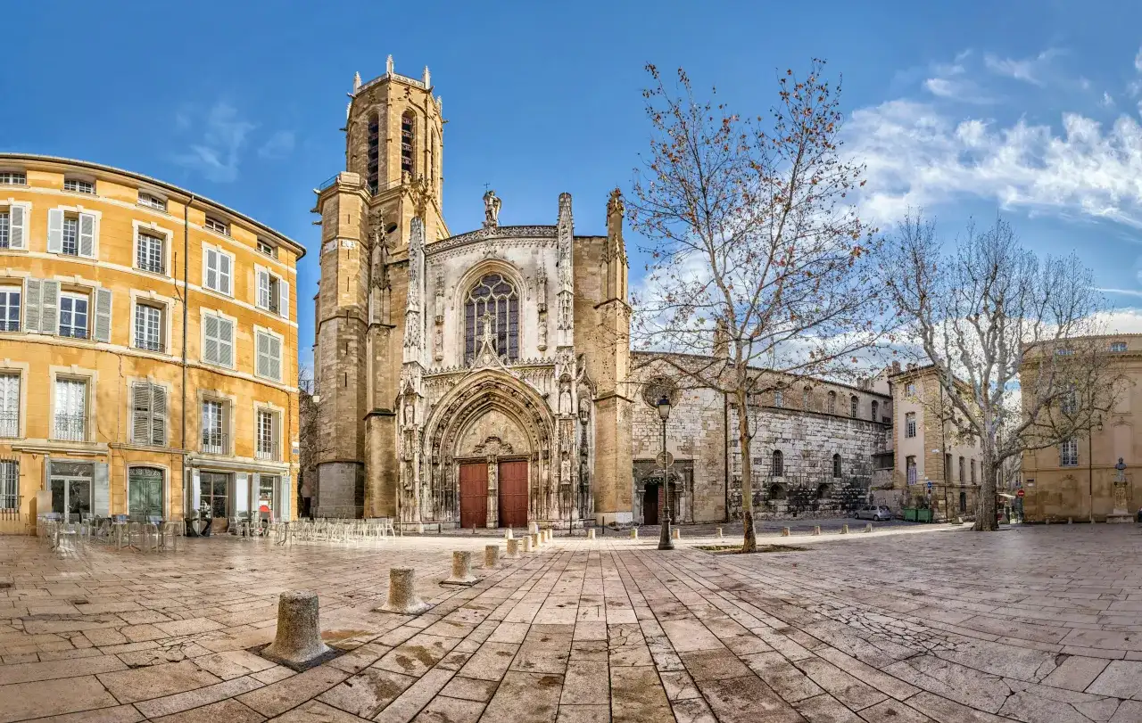 Katedra w Aix-en-Provence, z żółtą kamienicą obok, na brukowanym placu pod błękitnym niebem.