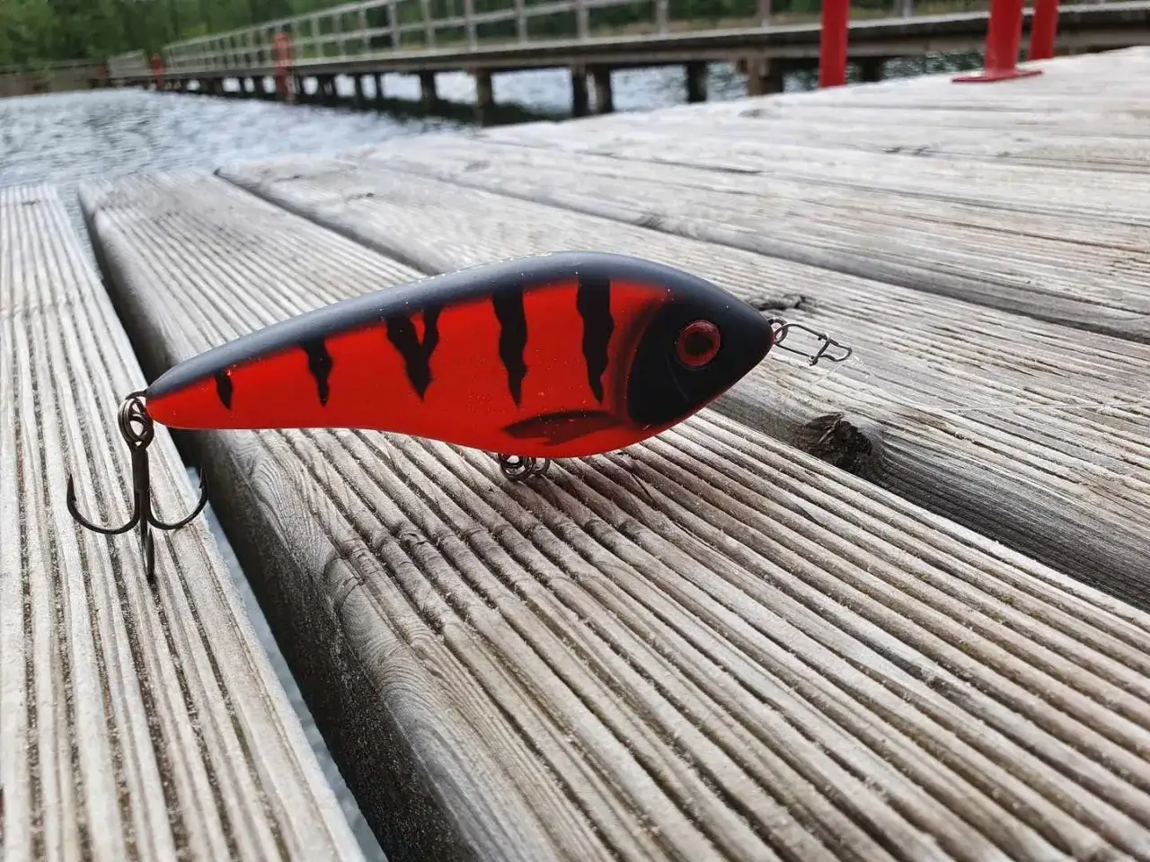 Czerwono-czarna przynęta wędkarska na szczupaka leżąca na drewnianym pomoście.