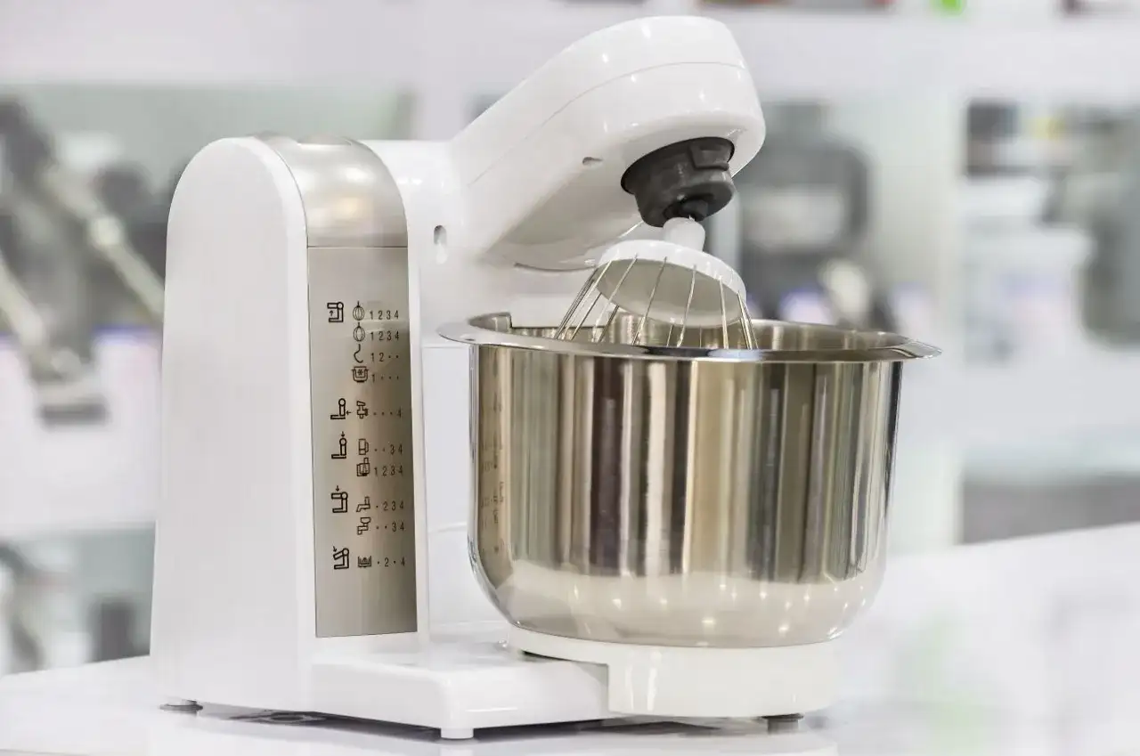 Biały robot kuchenny z metalową misą i trzepaczką. Na obudowie widoczne ikony funkcji, pomocne przy wyborze, jaki robot kuchenny wybrać blog.