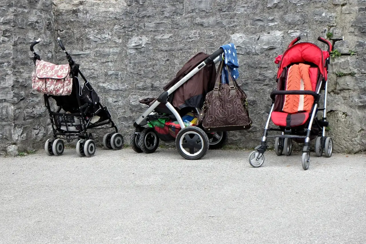 Trzy puste wózki dziecięce stoją przy kamiennej ścianie. Jeden ma torbę w kwiaty, drugi brązową torbę i niebieską chustę, a trzeci pomarańczowy kocyk.