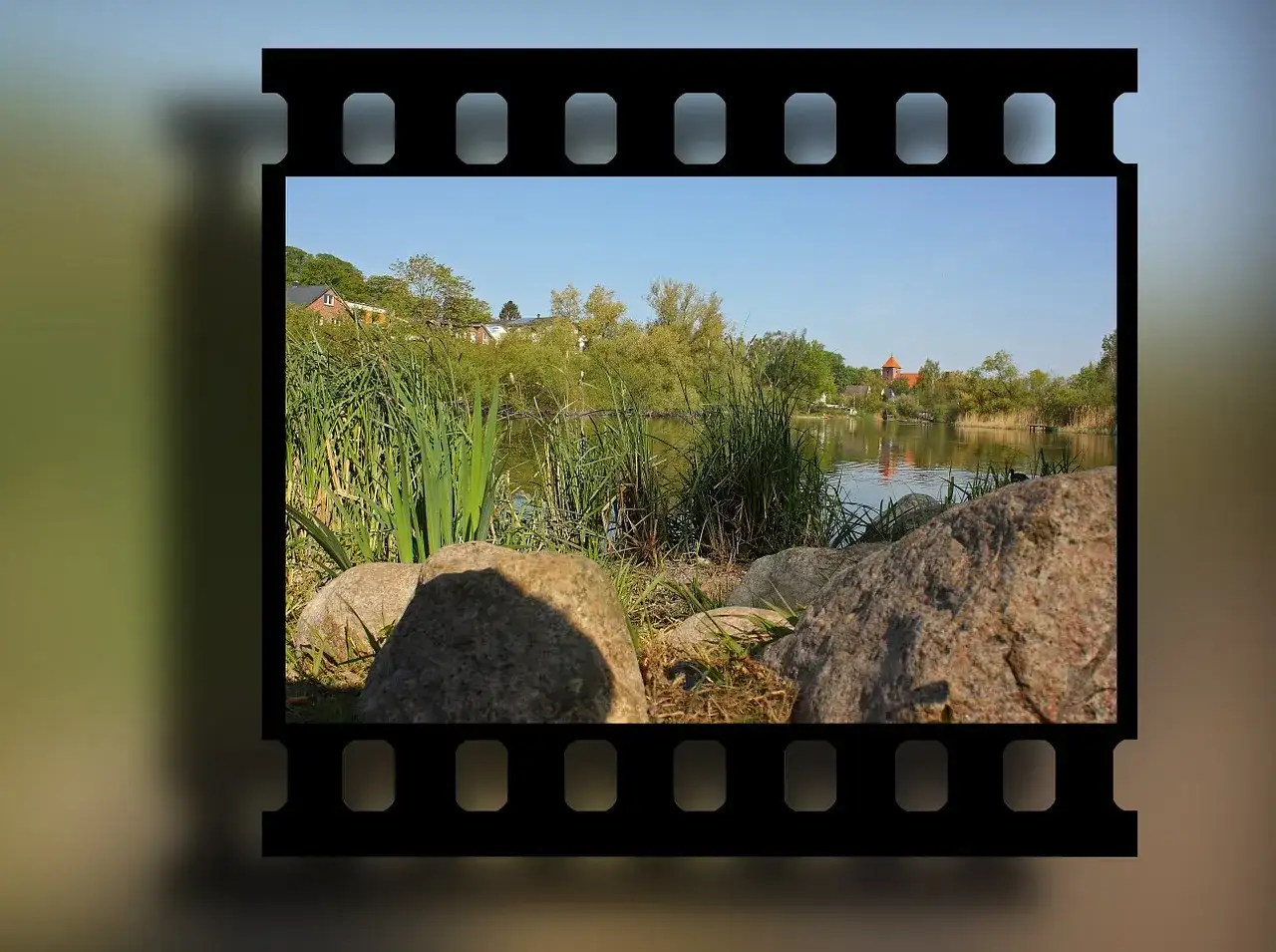 Obraz na kliszy fotograficznej: spokojny widok na jezioro z trzcinami, kamieniami i domami w tle.