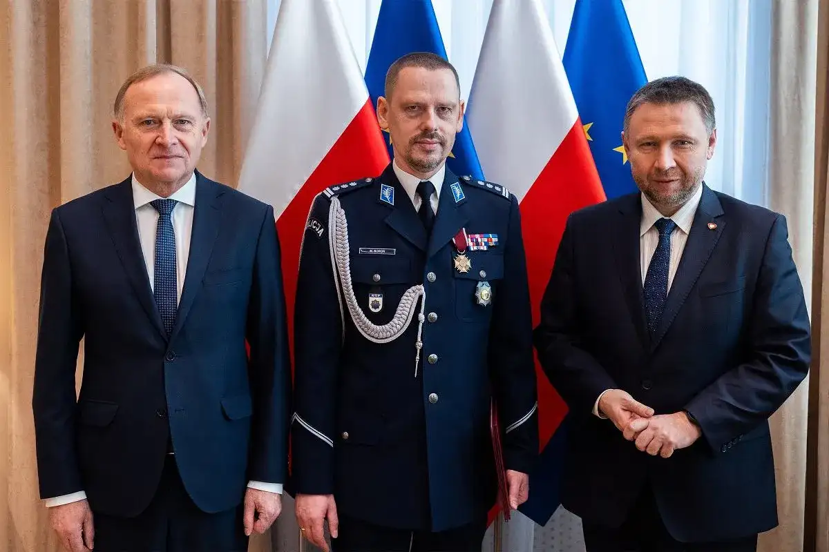 Trzech mężczyzn w garniturach i mundurze policyjnym stoi przed flagami Polski i UE.