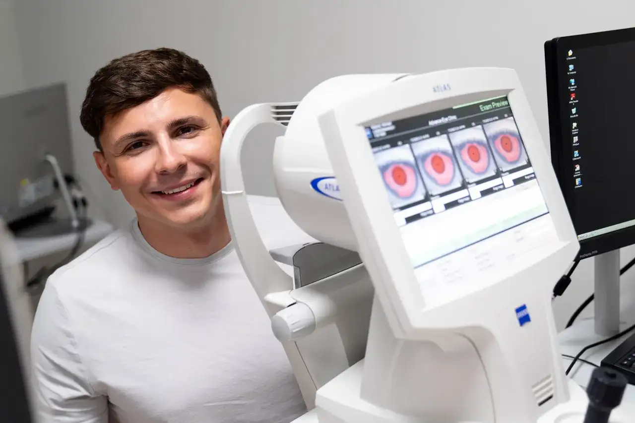 Uśmiechnięty mężczyzna obok urządzenia okulistycznego z obrazami oczu na ekranie.