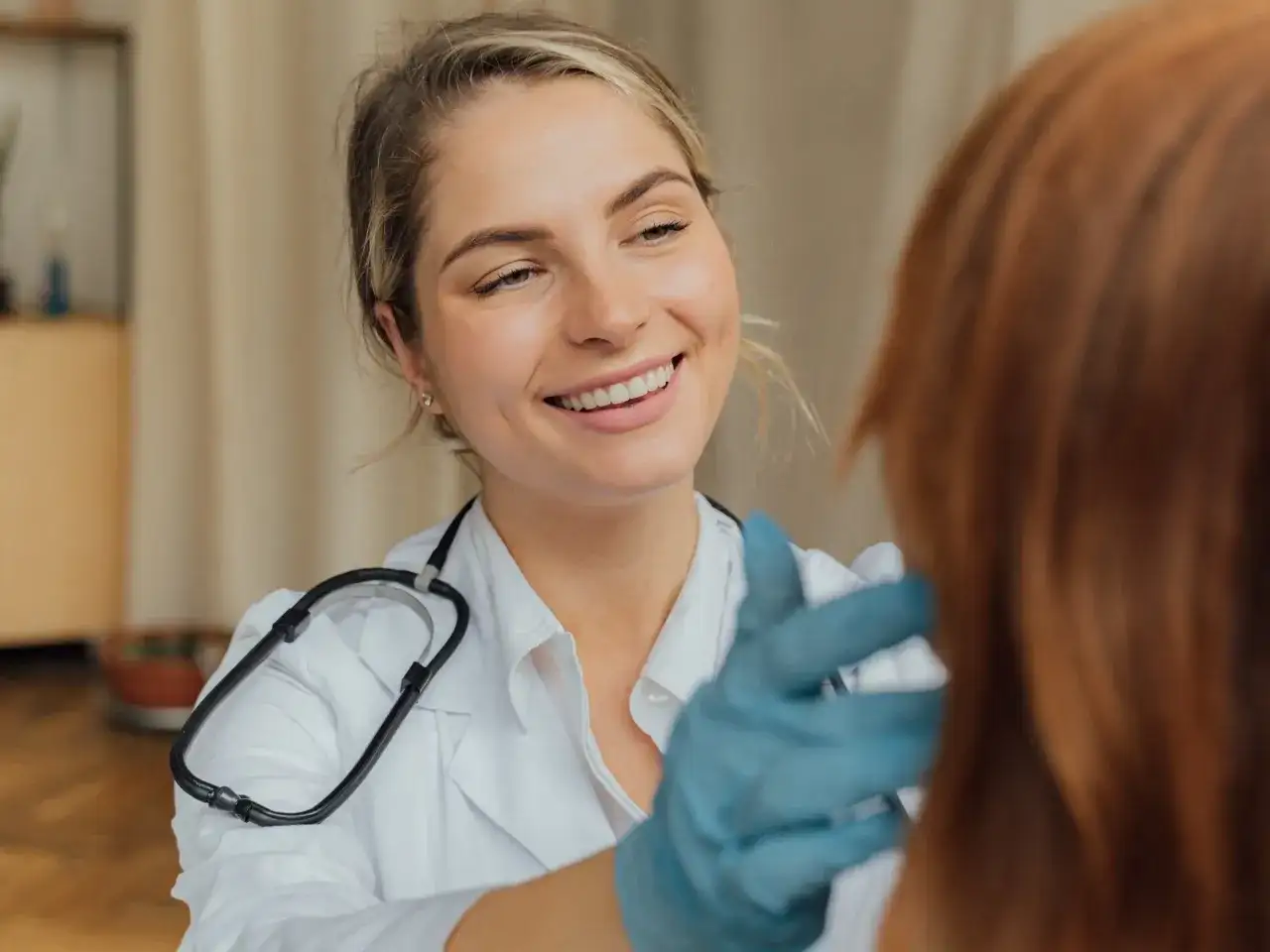 Uśmiechnięta lekarka w białym kitlu i niebieskich rękawiczkach bada pacjenta. Czy 16 latek może sam iść do lekarza? Tak, w wielu przypadkach jest to możliwe.