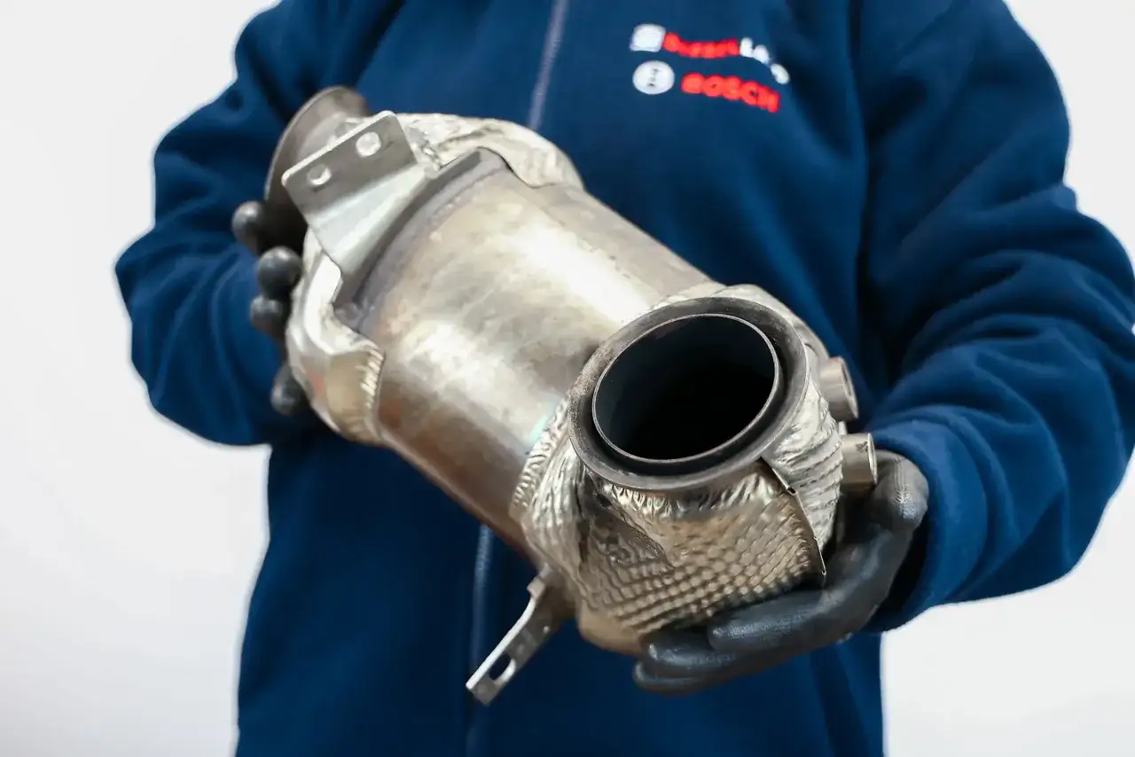 Osoba w niebieskiej bluzie i czarnych rękawiczkach trzyma metalowy element układu wydechowego.