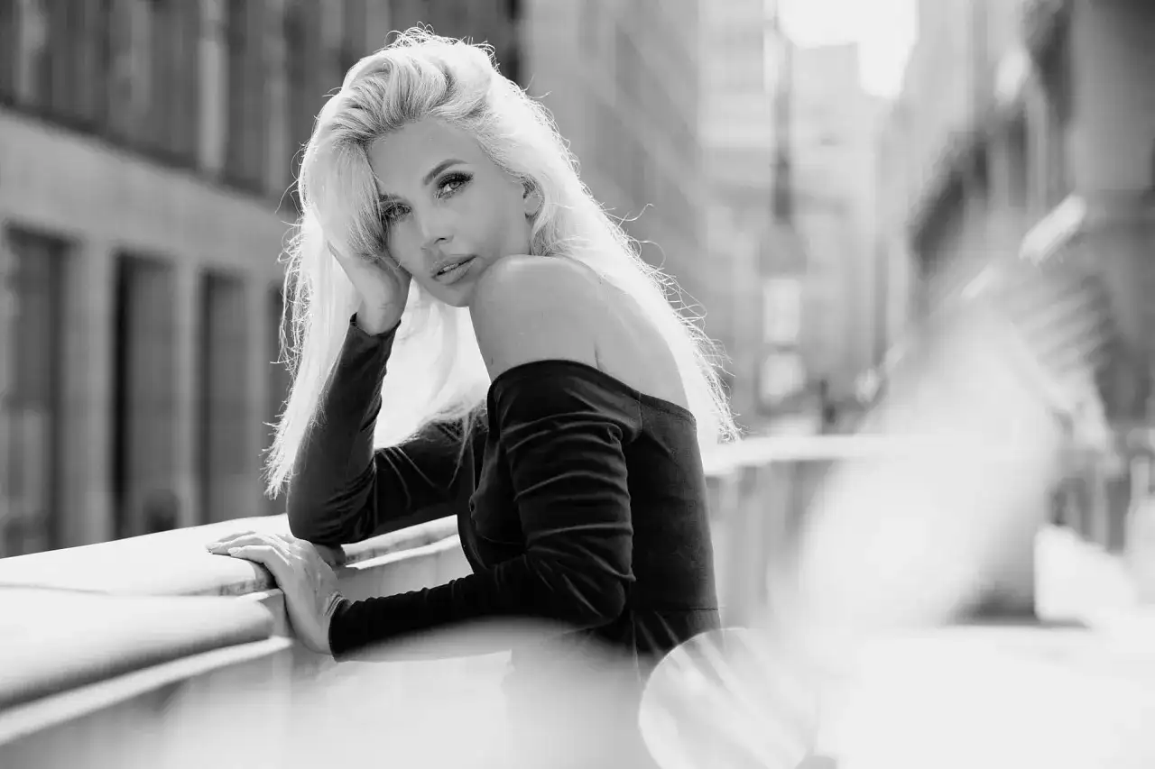 Elegancki portret w plenerze. Kobieta z blond włosami opiera się o balustradę, patrząc w obiektyw.