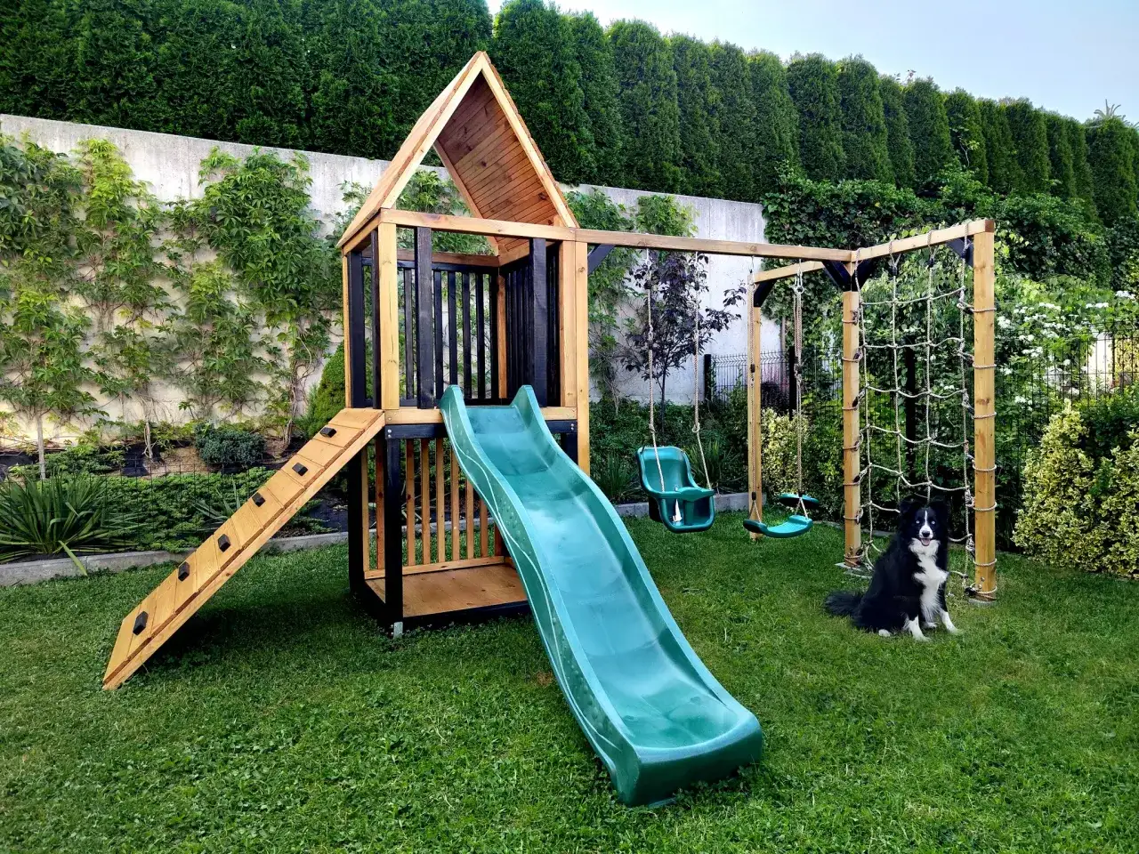 Drewniany plac zabaw z zieloną zjeżdżalnią, huśtawkami i ścianką wspinaczkową, na trawniku, z psem rasy border collie.