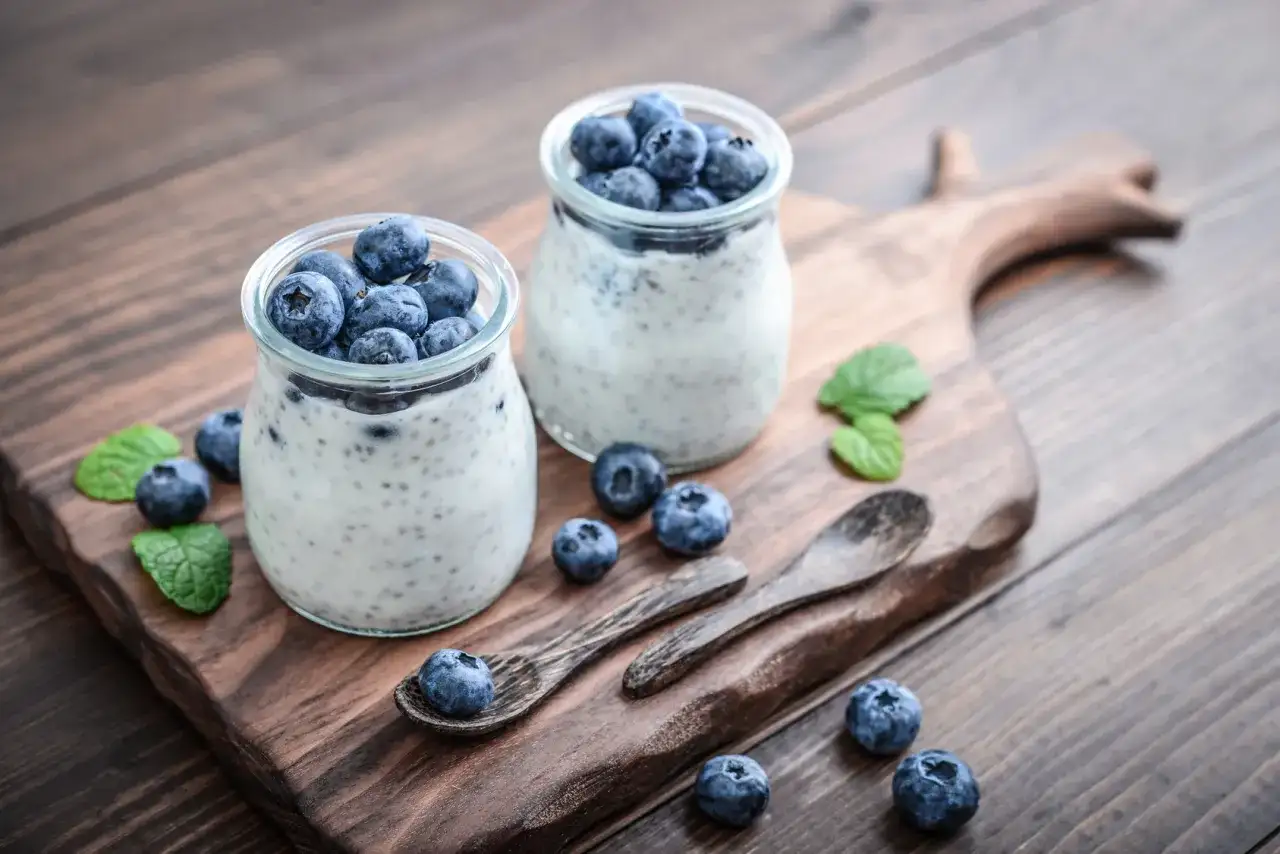 Dwa słoiczki z deserem pudding chia, ozdobione świeżymi borówkami i listkami mięty, na drewnianej desce.