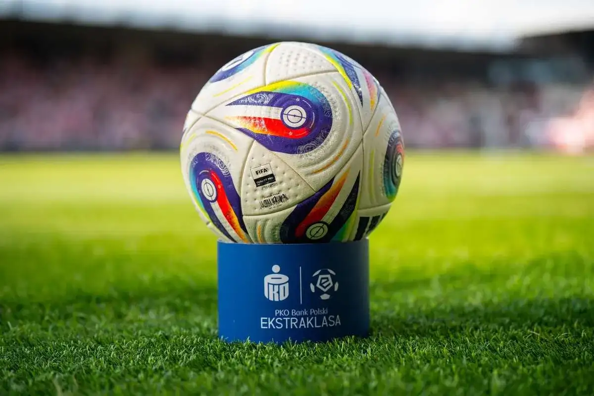 Piłka do piłki nożnej na niebieskim postumencie z logo Ekstraklasy na tle stadionu.
