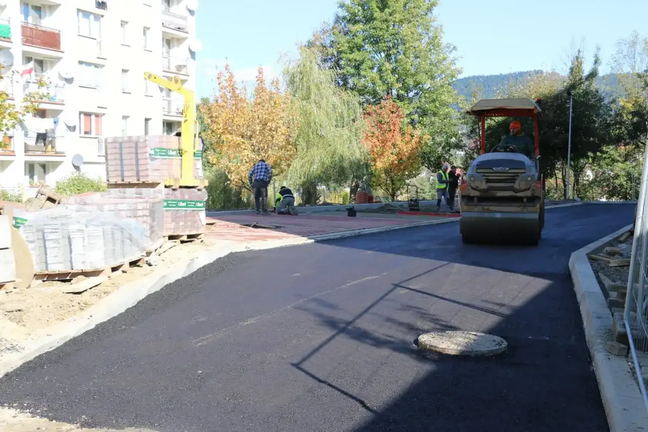 Prace drogowe na osiedlu Zygmunta Augusta w Limanowej. Walec drogowy ubija asfalt, a pracownicy układają kostkę.