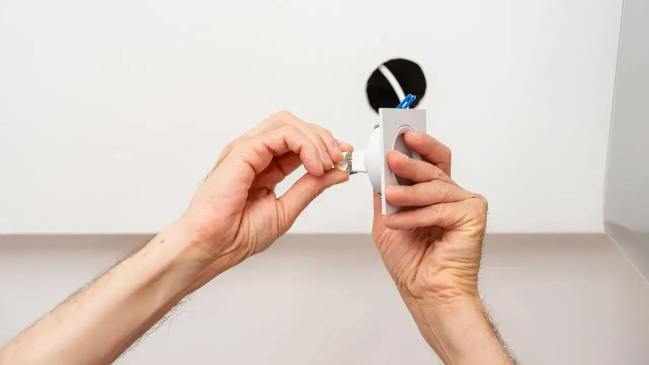 Dłonie montują oprawę lampy sufitowej, przygotowując ją do podłączenia. Widać tu fragmenty przewodów i otwór w suficie, co sugeruje instalację. Pytanie, jakie kołki do lampy sufitowej, jest kluczowe dla stabilnego montażu.