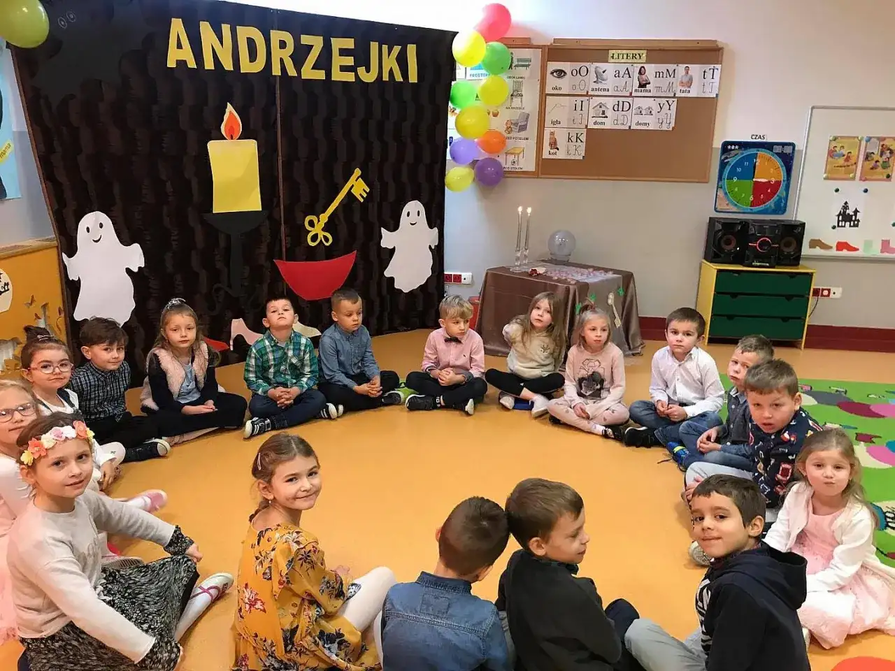 Dzieci na andrzejkach w przedszkolu, siedzą w kółku. W tle dekoracje z napisem "ANDRZEJKI", świece i duchy.