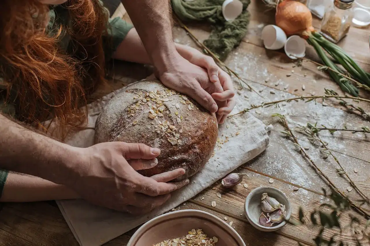 Dłonie obejmują bochenek chleba, jakby zastanawiając się, dlaczego chleb nie wyrasta. Na drewnianym stole mąka, jajka, cebula i czosnek.