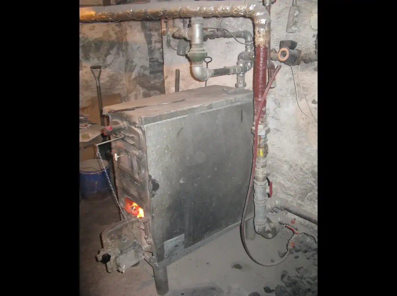 Stary piec węglowy z płomieniami w palenisku, otoczony rurami i narzędziami. Trudno ocenić, ile waży piec węglowy, ale jest solidny.