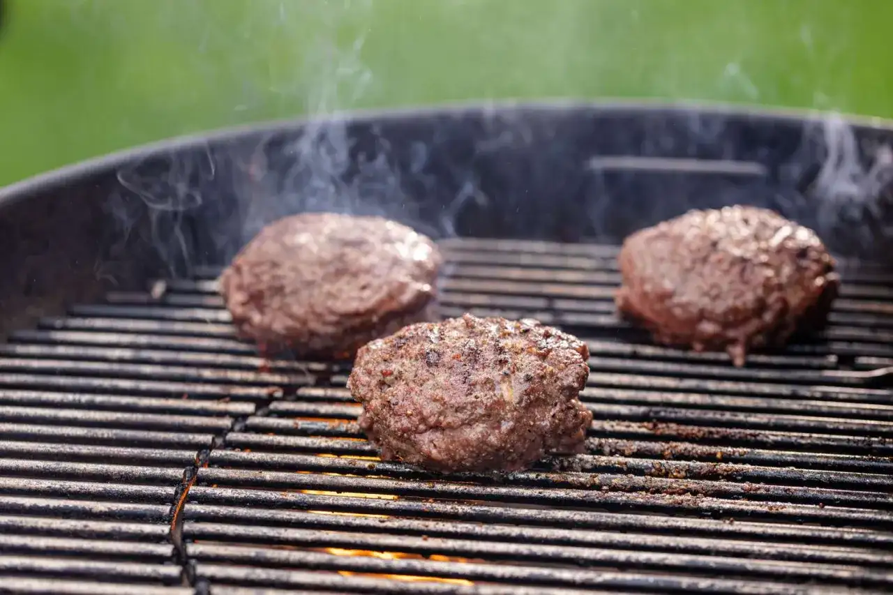 Trzy soczyste kotlety do burgerów smażące się na grillu, z których unosi się dym.