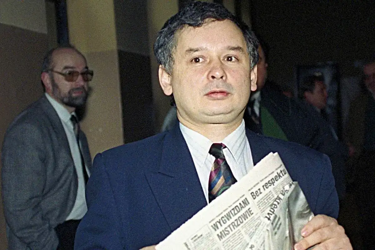 Jarosław Kaczyński, jeden z tych, kto założył PiS, trzyma gazetę. Obok stoi mężczyzna w okularach.