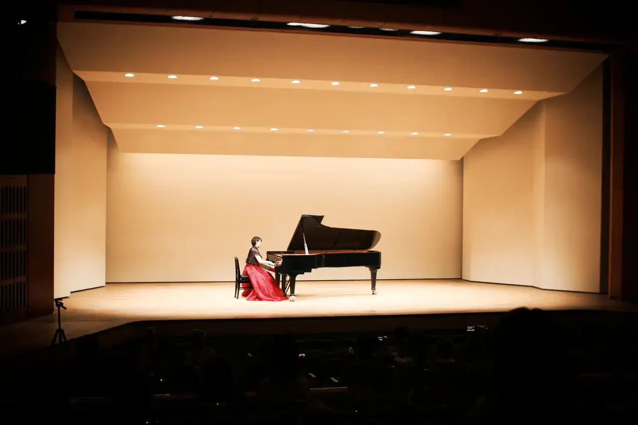 Kobieta w czerwonej sukni gra na fortepianie podczas kameralnego recitalu. Publiczność w ciemnościach czeka na występ.