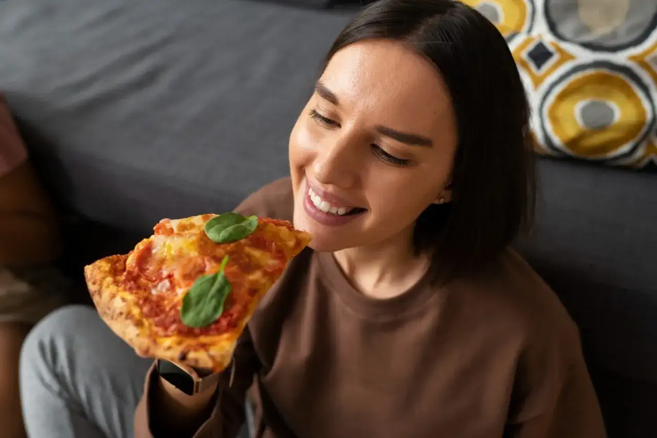 Uśmiechnięta kobieta delektuje się kawałkiem pizzy, zastanawiając się, jak podgrzewać pizzę, by była idealna.